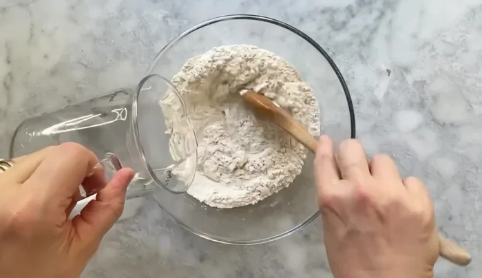 Preparare l'impasto: In una ciotola, mescolare la farina e il lievito secco (47.52). Aggiungere gradualmente l'acqua, tenendone da parte un po' (51.62). Impastare con un cucchiaio, poi con le mani fino ad ottenere un composto omogeneo (59.6). Aggiungere il sale sciolto in poca acqua e l'olio (71.84, 86.13). Lavorare l'impasto fino a renderlo liscio (79.63).