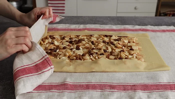 Arrotolare lo strudel: Piegare i lati lunghi della pasta e arrotolare partendo da un lato corto, aiutandovi con il canovaccio. Trasferire sulla teglia foderata con carta forno, con la chiusura rivolta verso il basso.